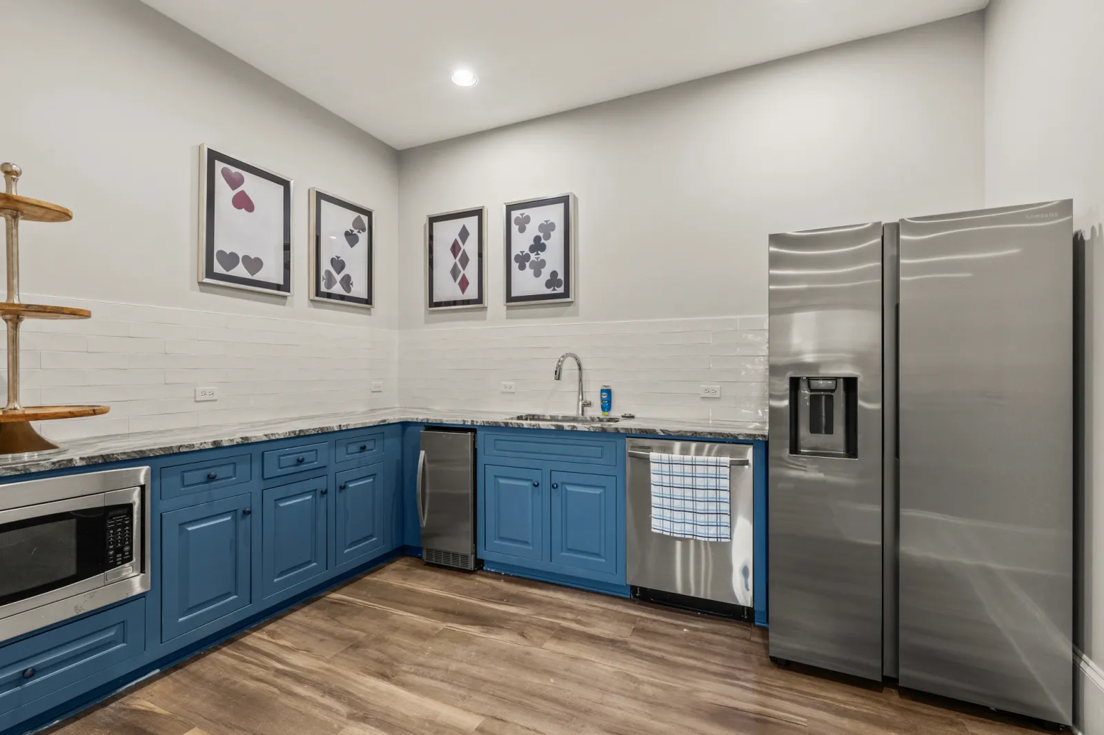 Modern kitchen with blue cabinets, stainless steel appliances, wooden flooring, and playing card art on white walls.