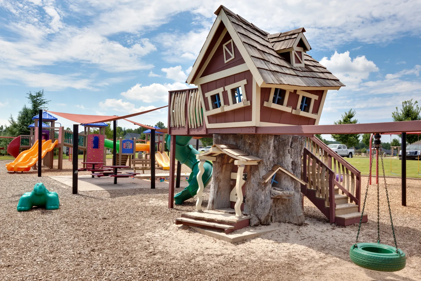 Playground with a whimsical treehouse, slides, swings, and colorful play structures under a blue sky.