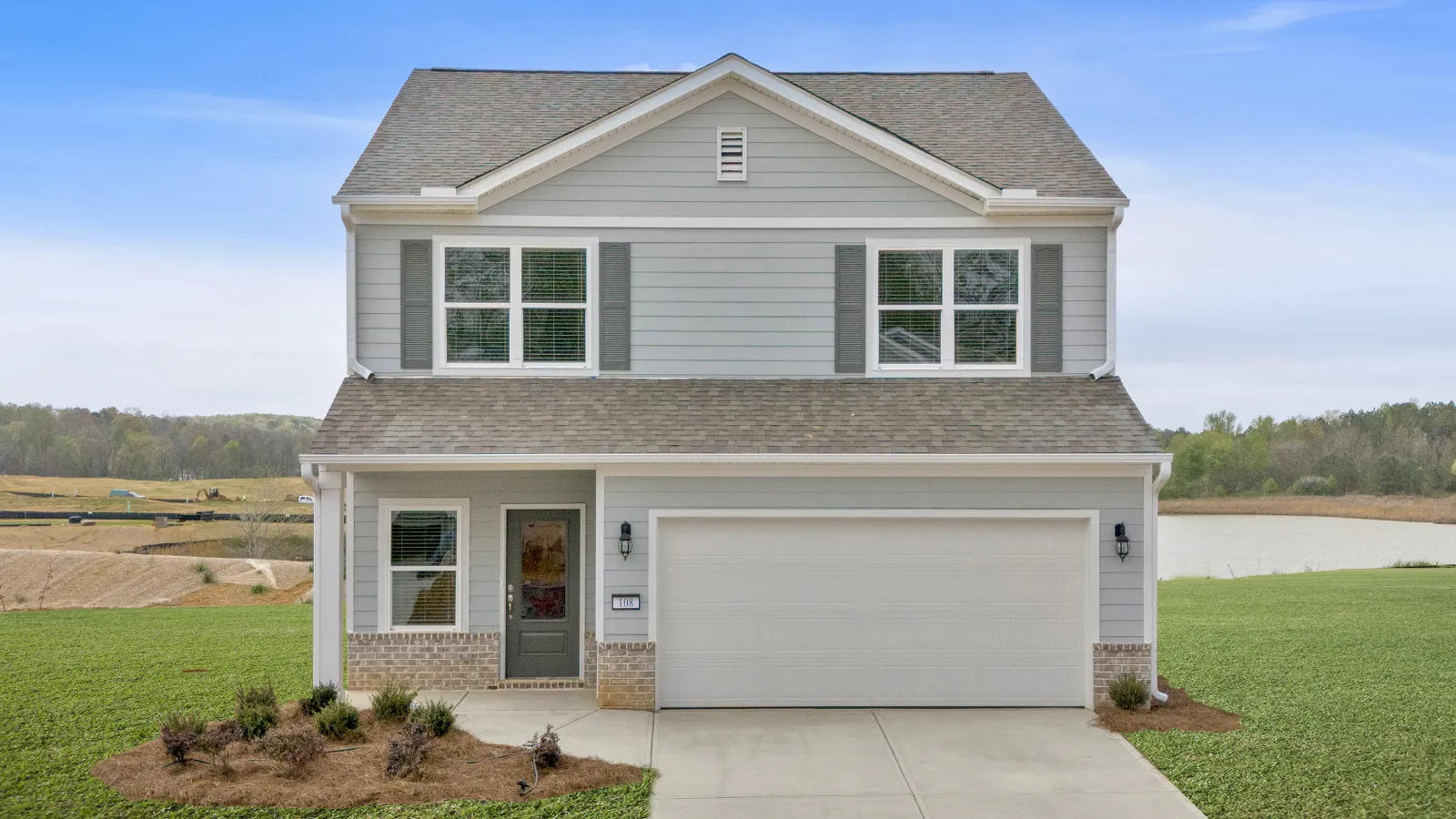 Two-story gray siding house with white trim, attached garage, landscaped front yard, and a pond in the background