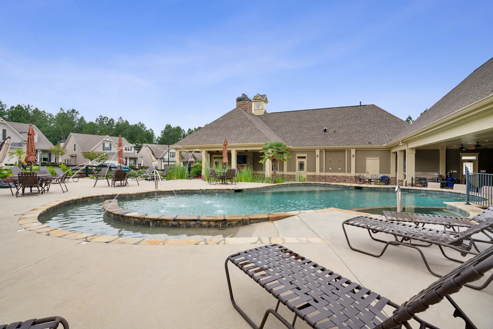 Community pool area with lounge chairs, tables, umbrellas, and residential houses in the background under blue sky.