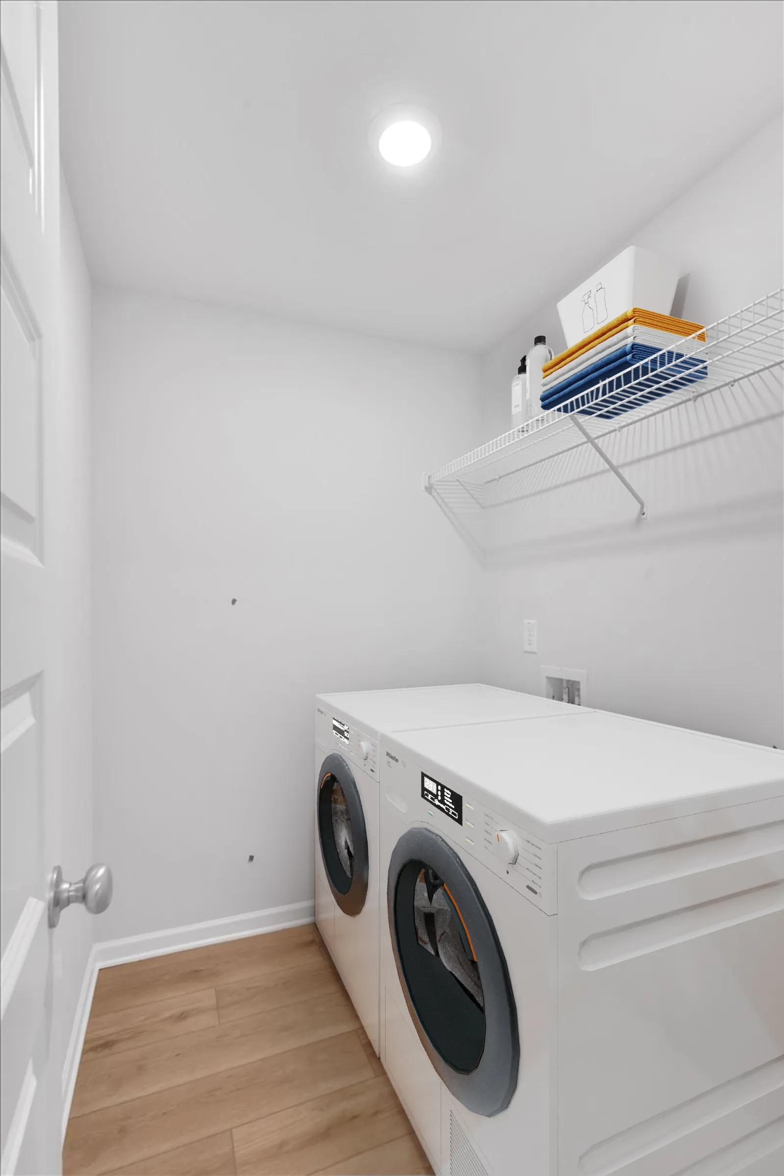 Bright laundry room with front-load washer and dryer, white walls, wire shelf with folded towels and detergent bottles.