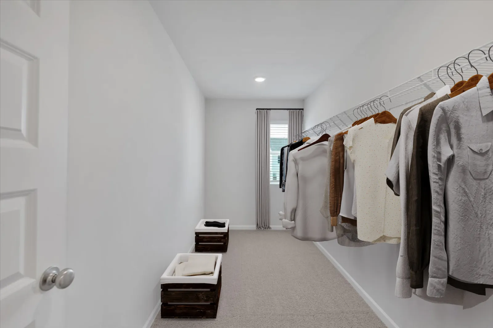 Minimalist walk-in closet with hanging clothes on wire rack, white walls, carpet floor, and window at the end.