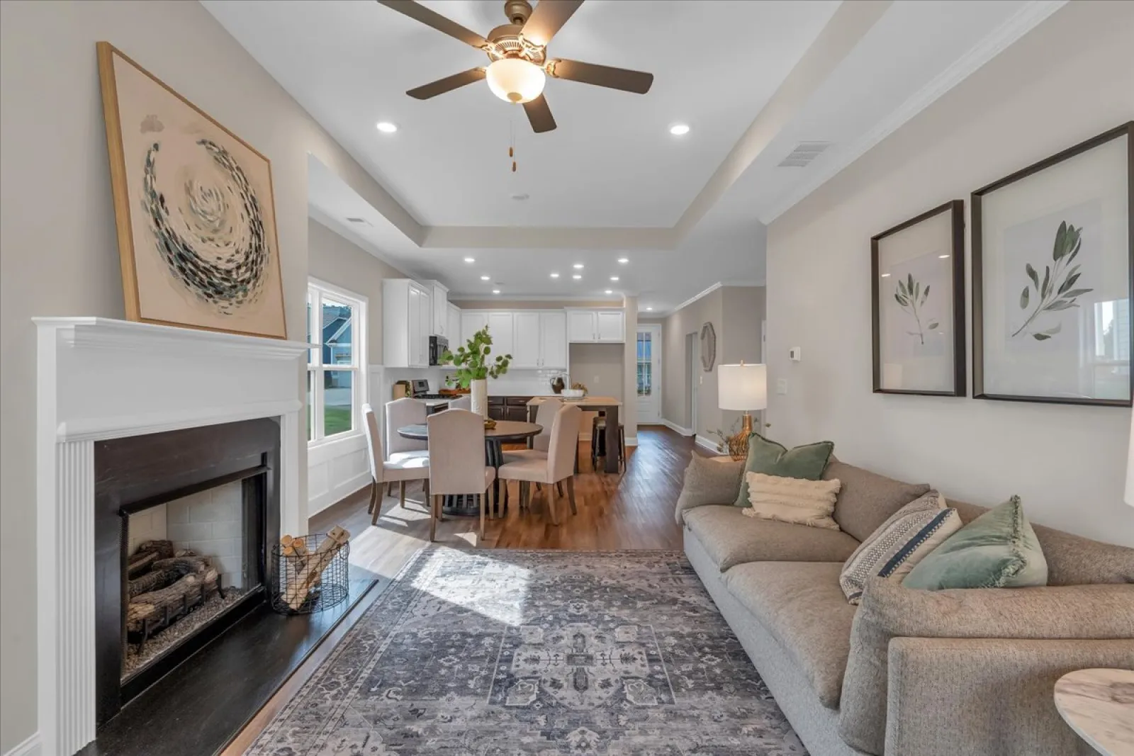 Bright open living room with fireplace, ceiling fan, gray sofa, dining table, and hardwood flooring under natural light