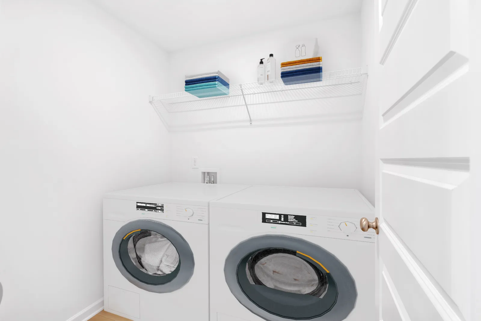 White laundry room with front-loading washer and dryer, wire shelf holding towels and bottles above appliances.