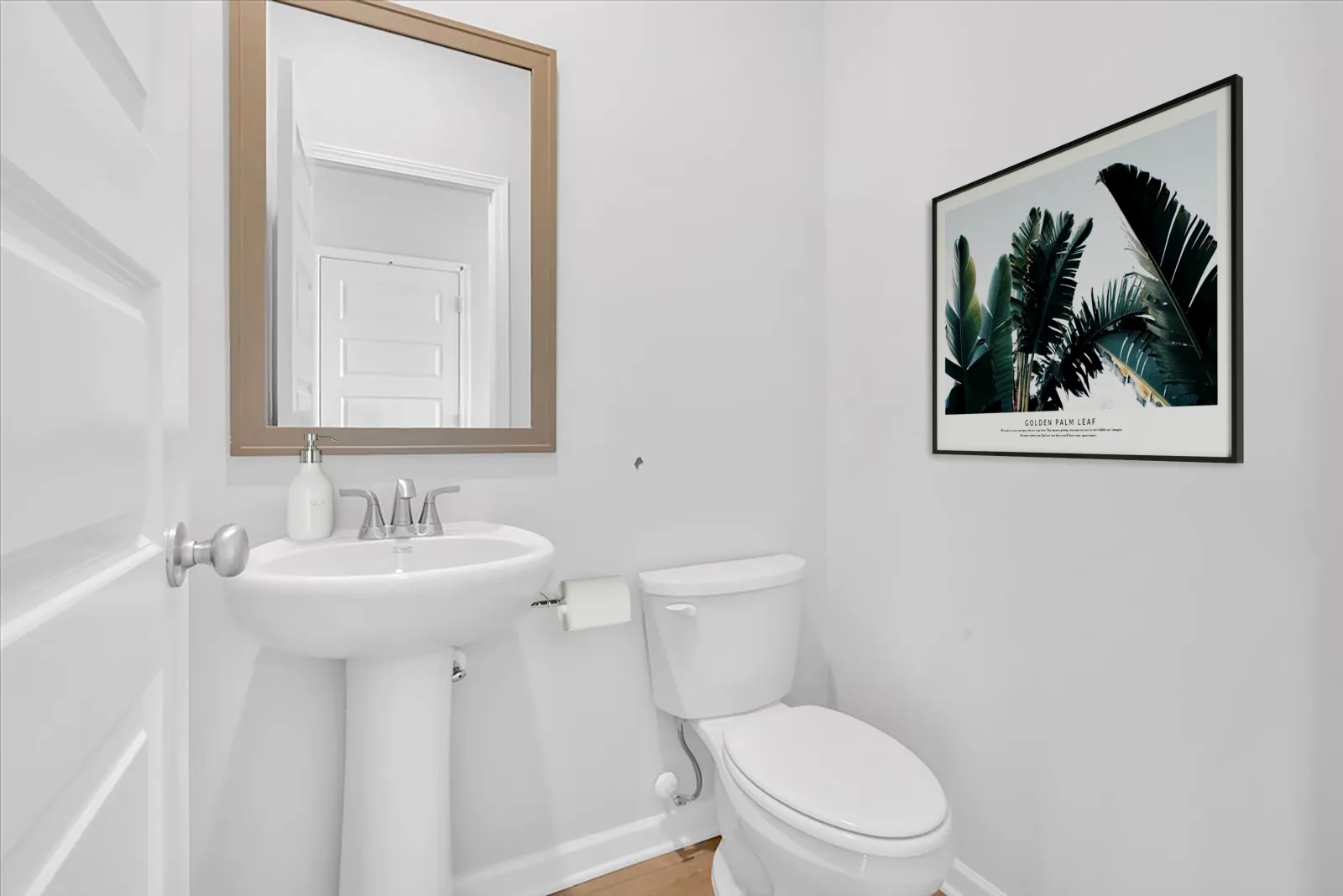 Small white bathroom with pedestal sink, toilet, mirror, and framed tropical leaf artwork on wall.