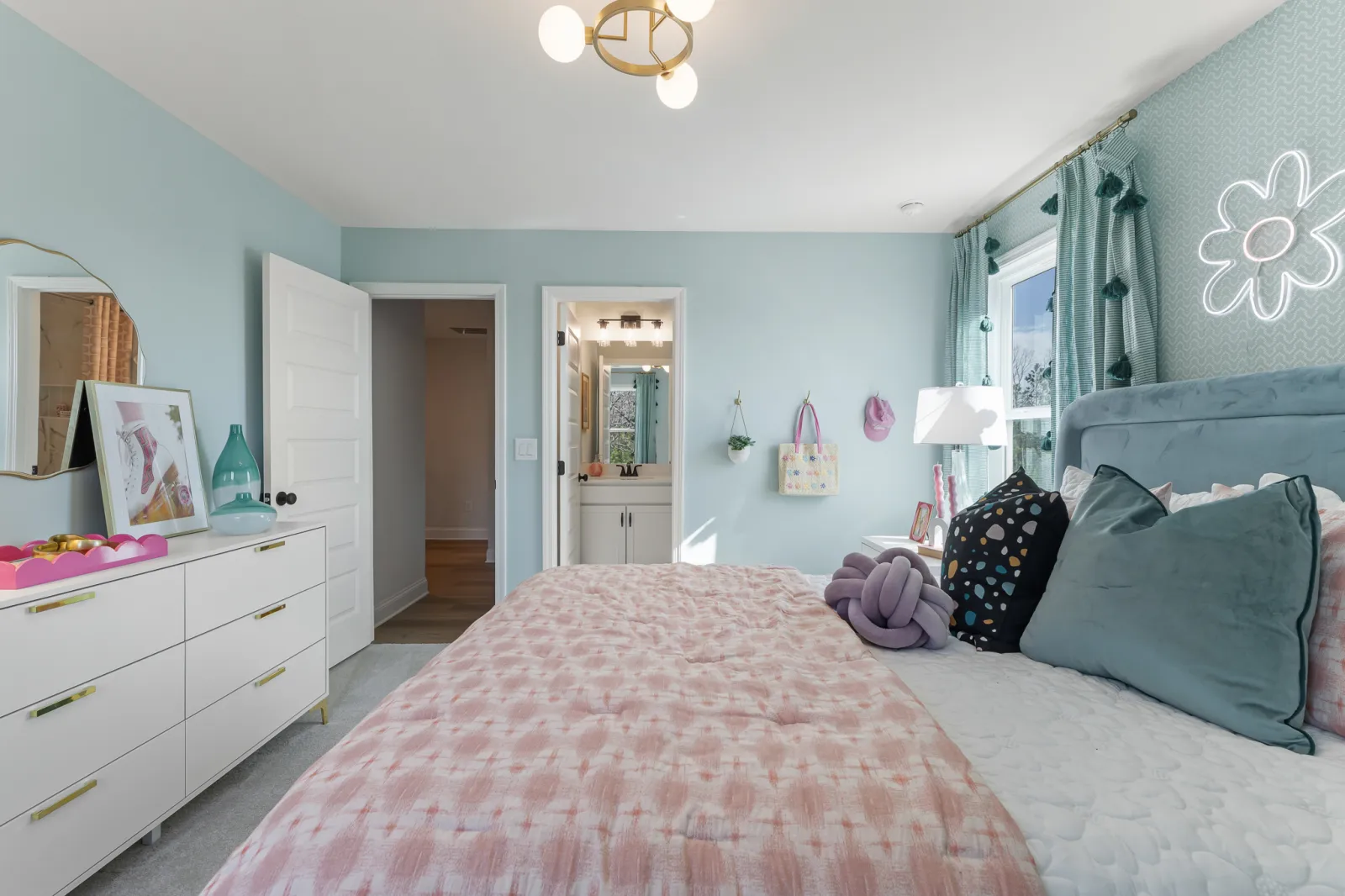 Bright bedroom with pastel decor, pink bedspread, teal pillows, white dresser, and a neon flower light on wall.