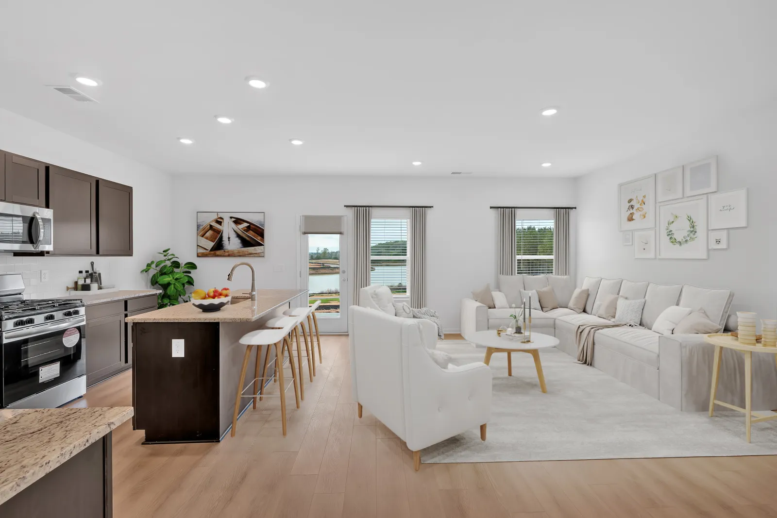 Modern open-concept living room and kitchen with white furniture, wood flooring, and large windows overlooking a lake.