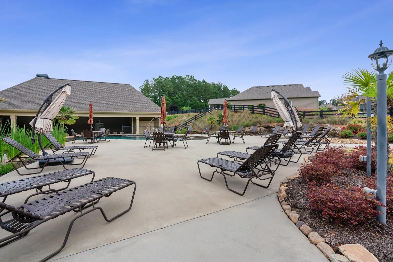 Outdoor poolside area with lounge chairs, tables, umbrellas, landscaped garden, and residential houses in background.