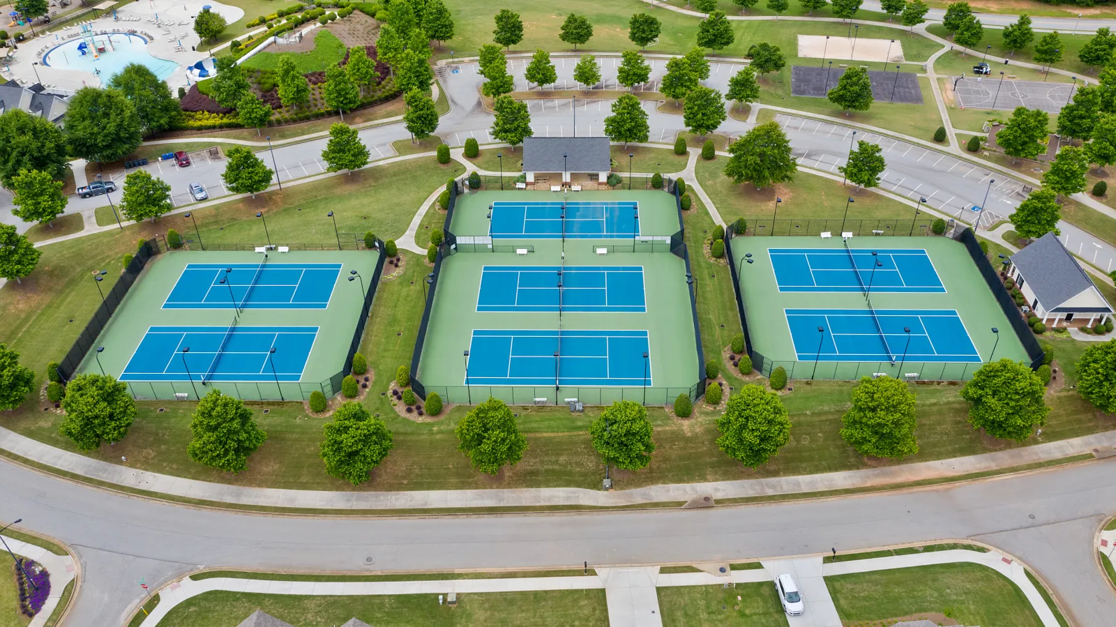 Aerial view of three fenced tennis courts with blue surfaces and green surroundings in a park area.