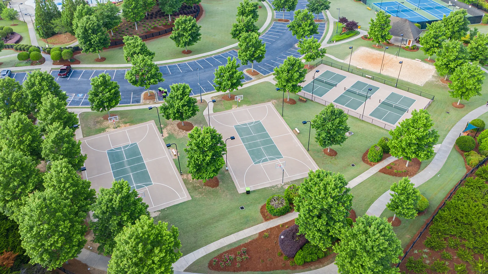 Aerial view of a park featuring two basketball courts, tennis courts, pathways, and lush green trees.