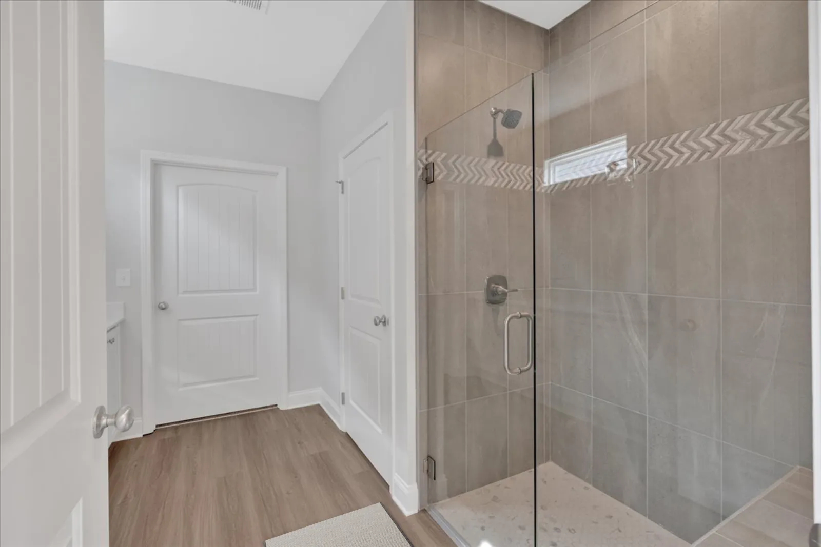 Modern bathroom with glass-enclosed shower, beige tiles, wooden floor, and white paneled doors.
