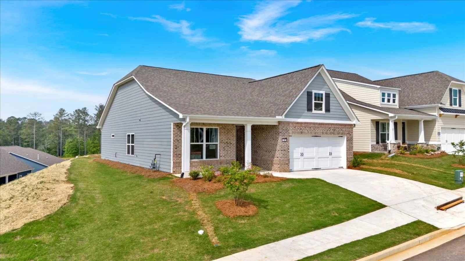 Modern single-story house with gray siding, brick facade, two-car garage, and landscaped front yard on a sunny day.