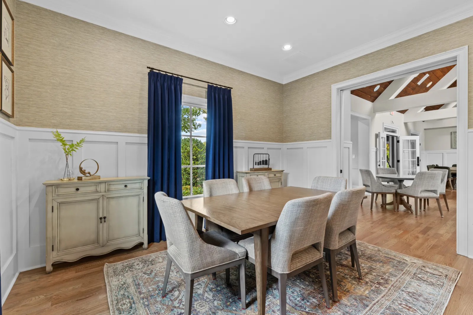 Modern dining room with six upholstered chairs, wooden table, blue curtains, and vintage sideboard on patterned rug.