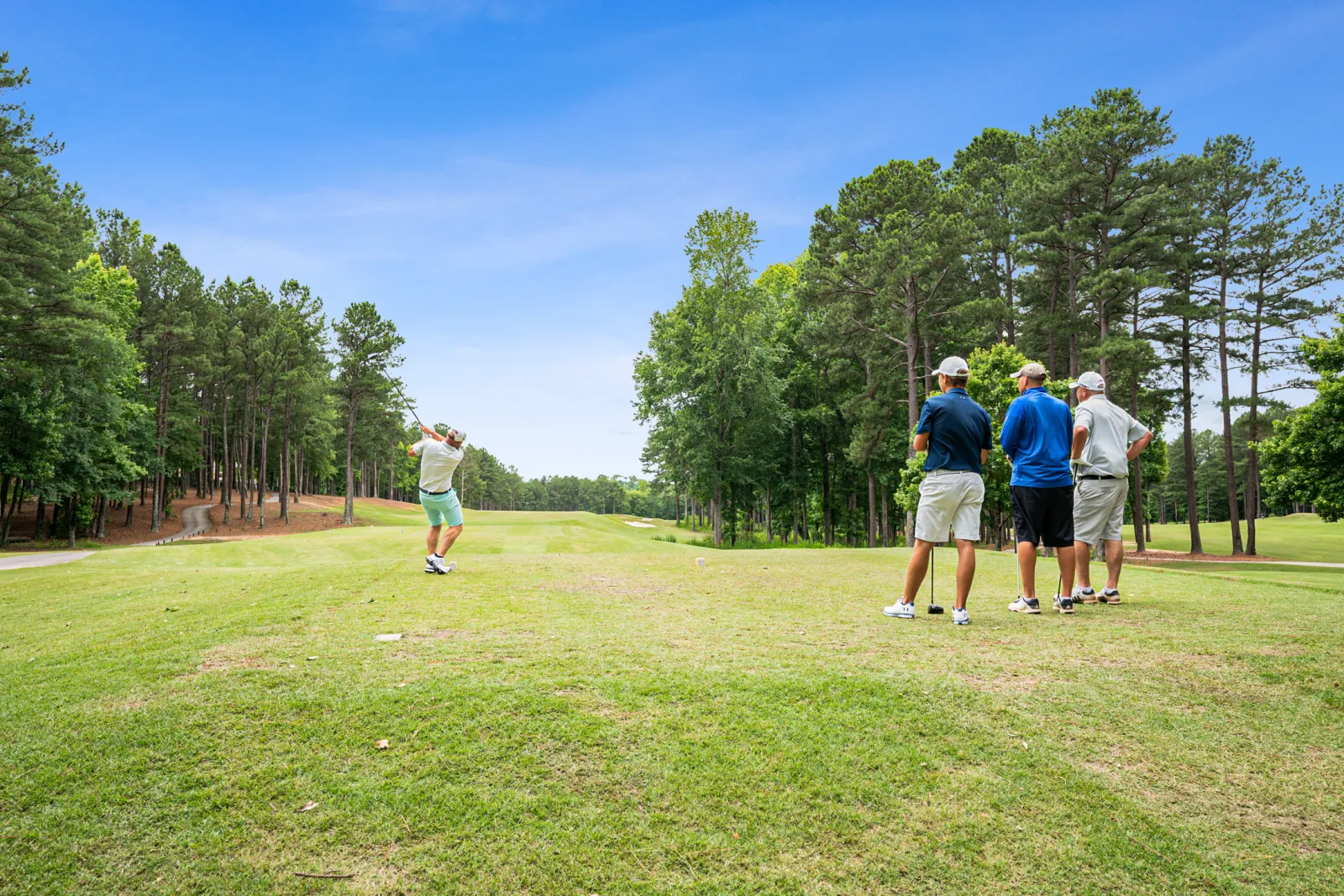 Four men play golf on a sunny day, one swinging a club while three watch on a green fairway surrounded by trees.