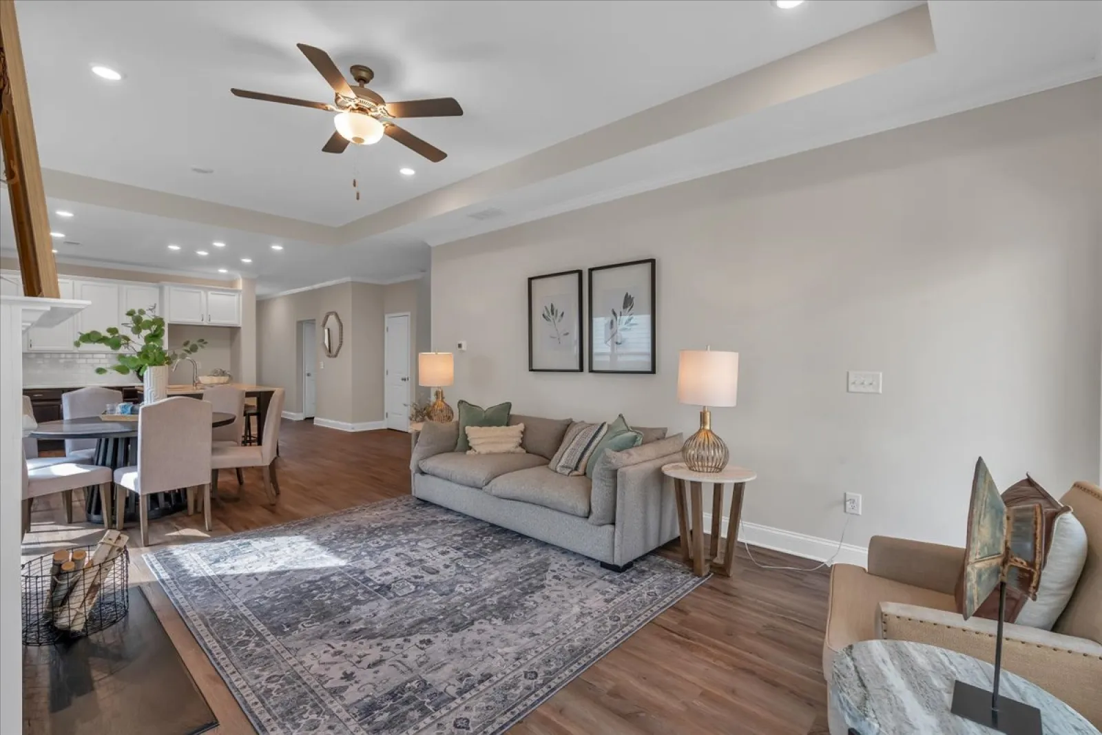 Modern living room with gray sofa, armchair, patterned rug, ceiling fan, and open dining area with hardwood floors.