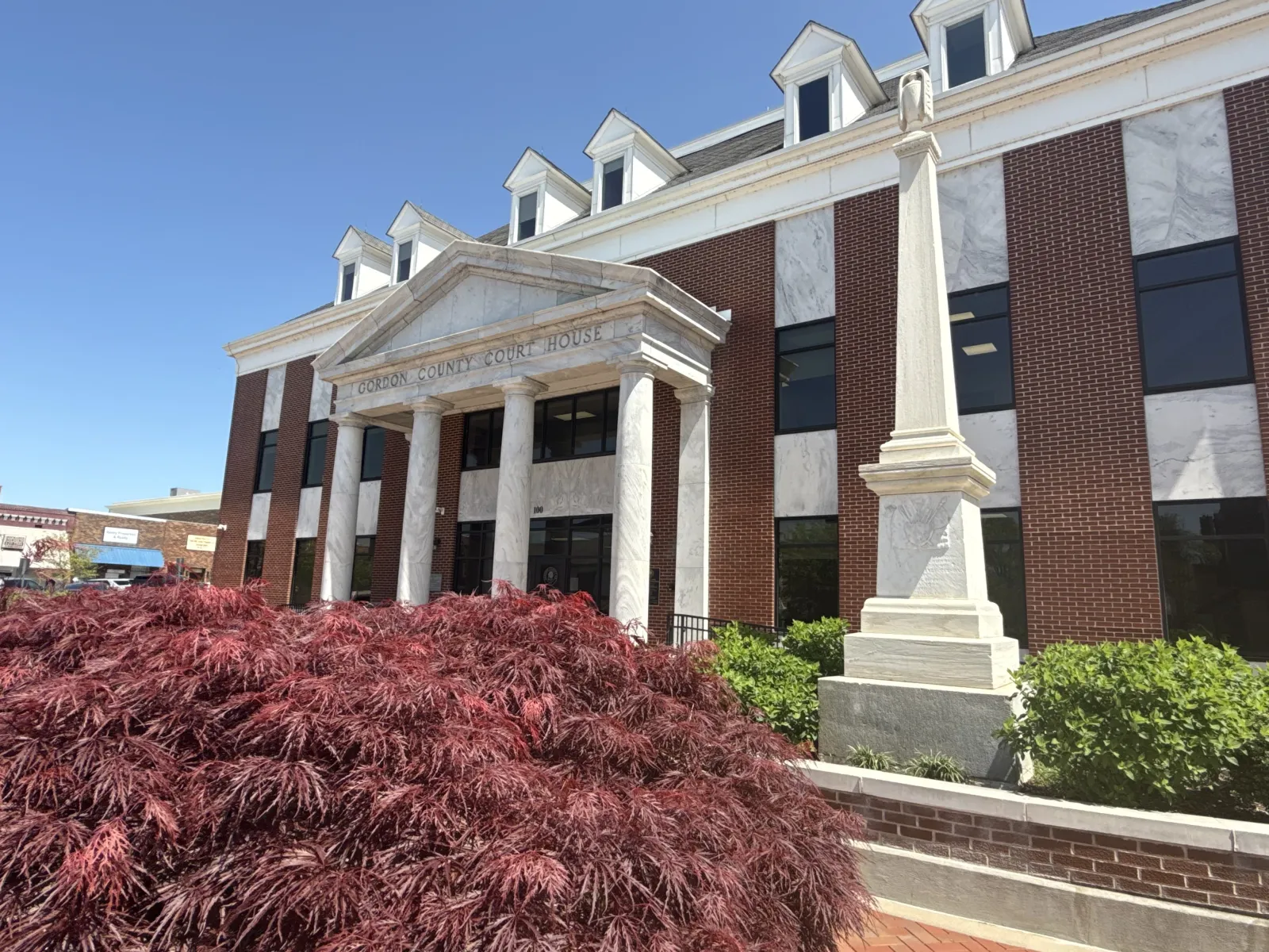 Gordon County Courthouse with white columns, red brick facade, obelisk monument, and red foliage under clear sky.