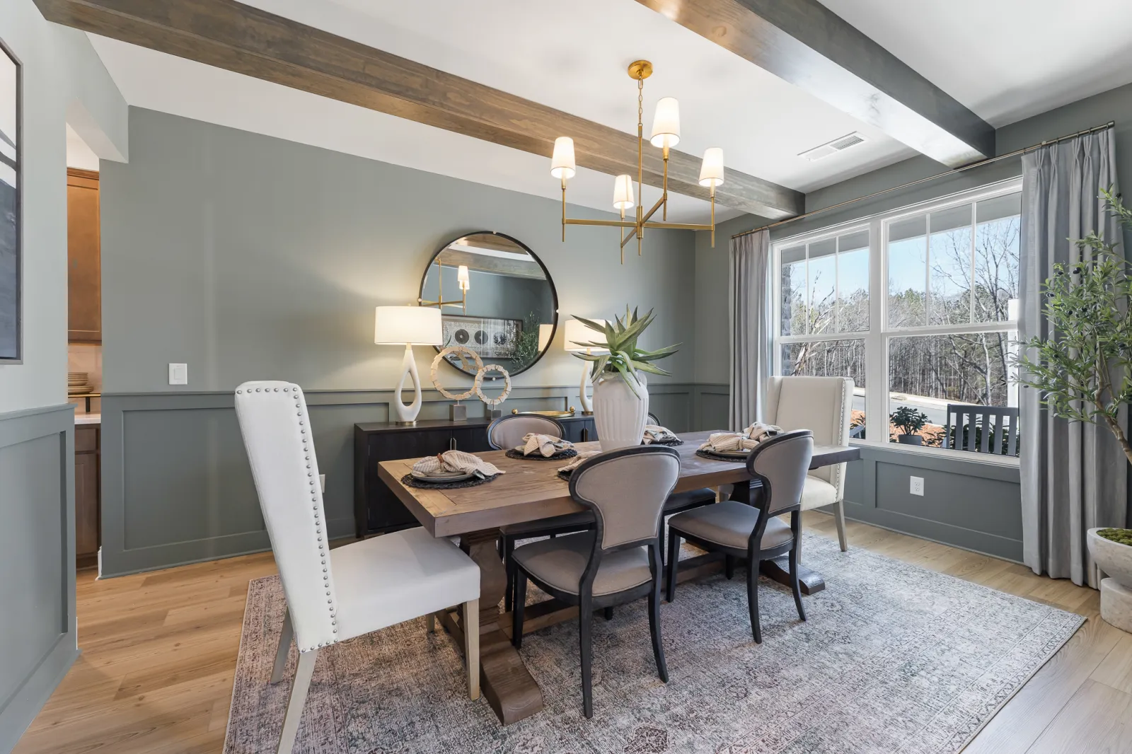 Modern dining room with wooden table, mixed chairs, round mirror, chandelier, large window, and potted plants.