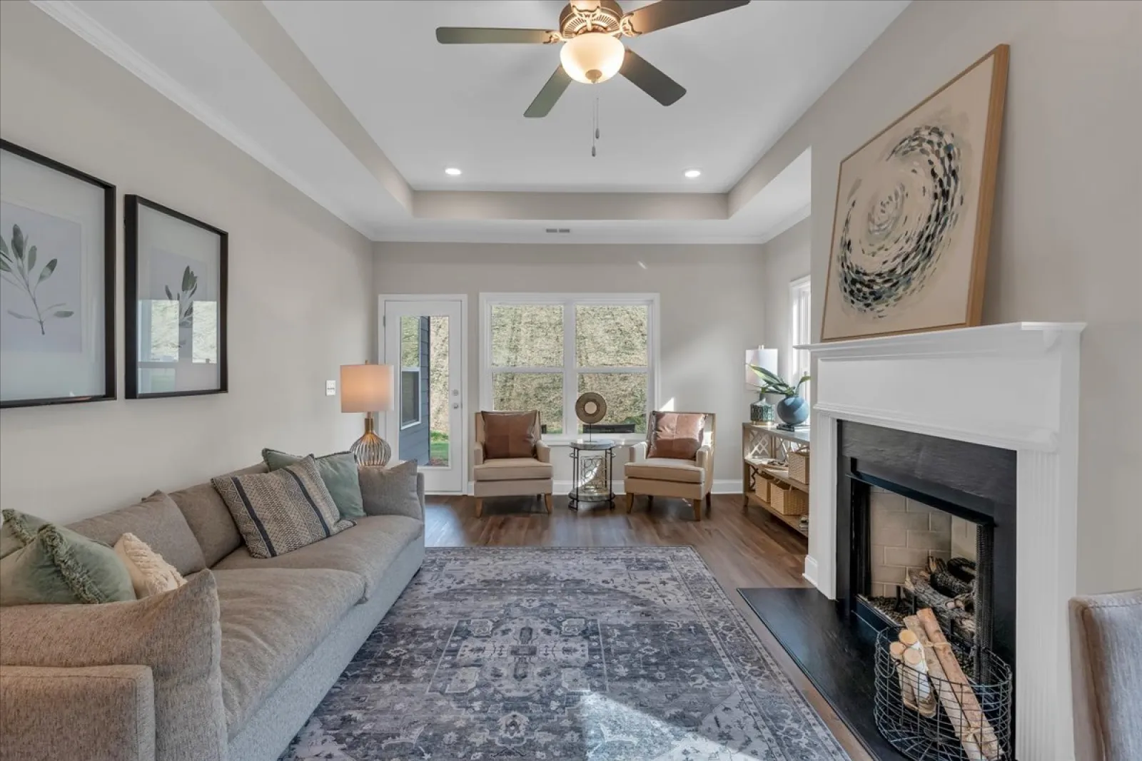 Bright living room with gray sofa, two armchairs, decorative rug, fireplace with art, and ceiling fan with light.