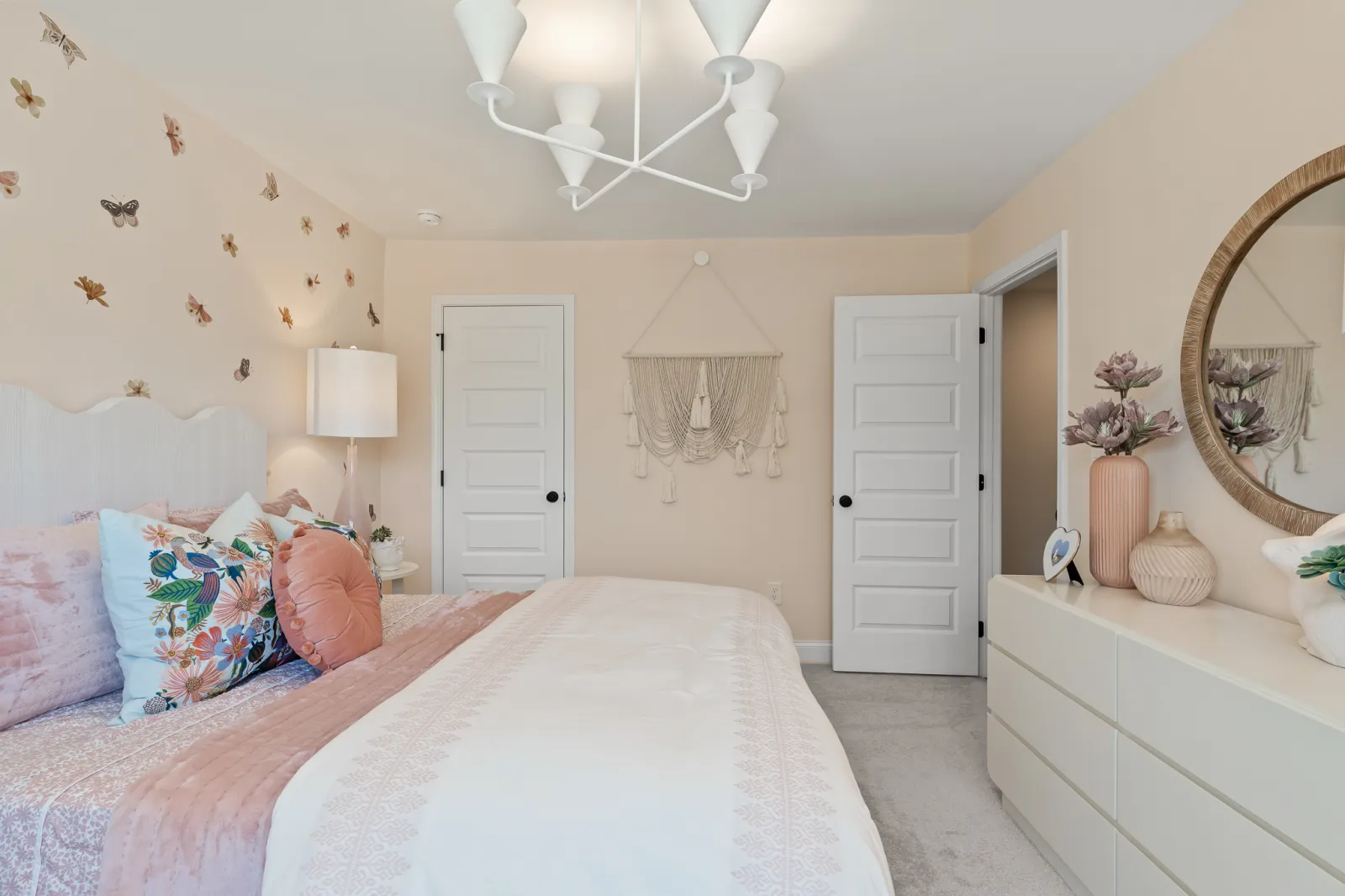 Cozy bedroom with floral pillows, butterfly wall decals, white dresser, round mirror, and soft pink and beige tones.