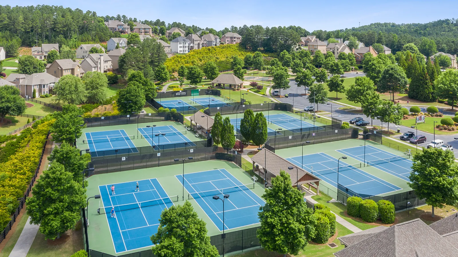 Aerial view of multiple tennis courts surrounded by trees and houses in a suburban neighborhood on a sunny day