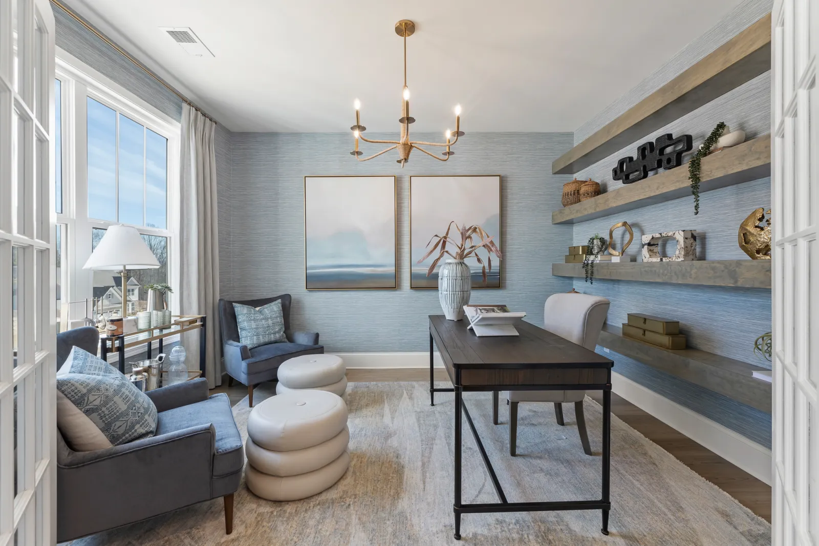 Modern home office with blue armchairs, wooden desk, floating shelves, soft rug, and large window light.