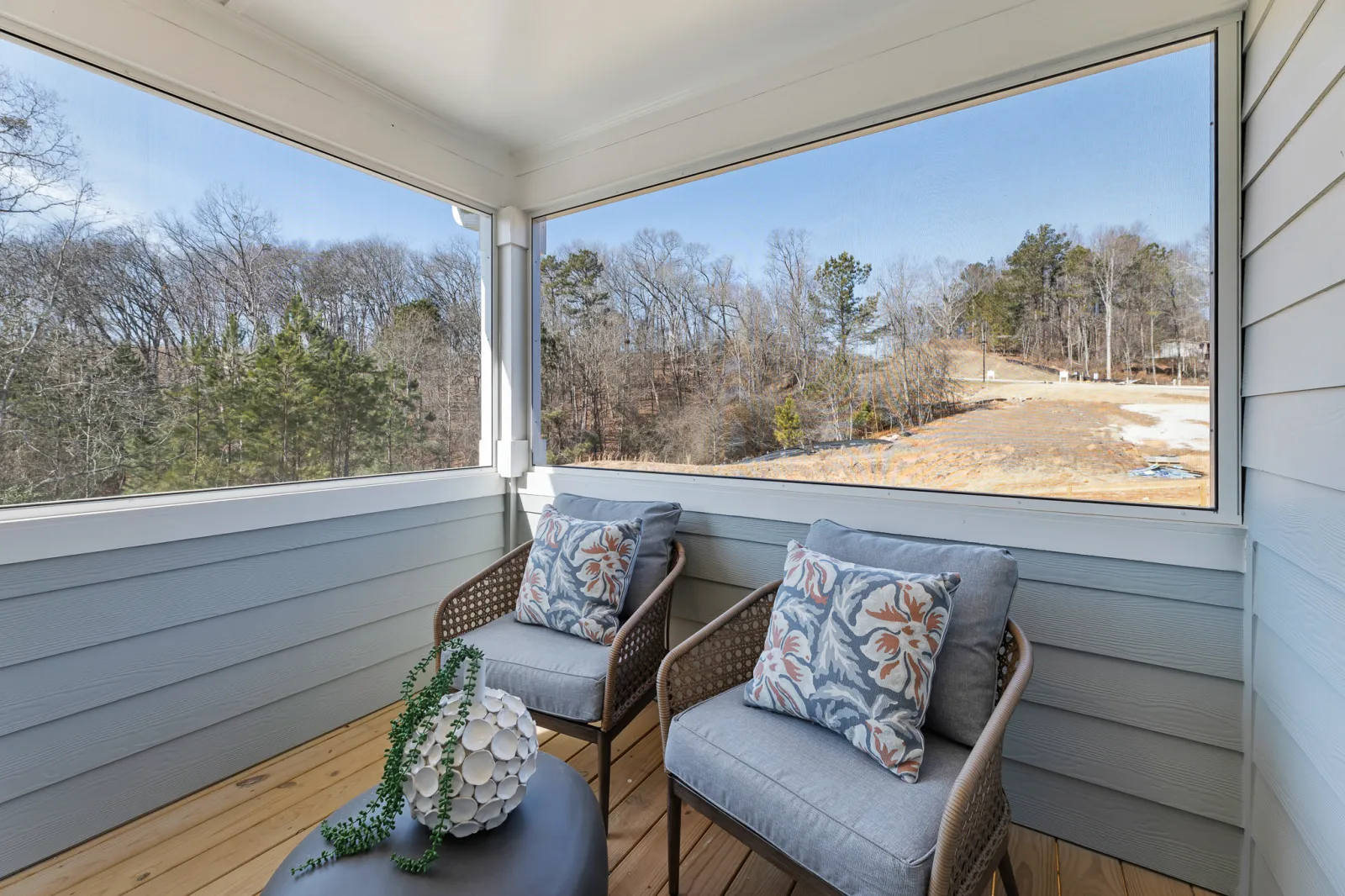 Cozy screened porch with two cushioned chairs, floral pillows, a small table, and a view of leafless trees.