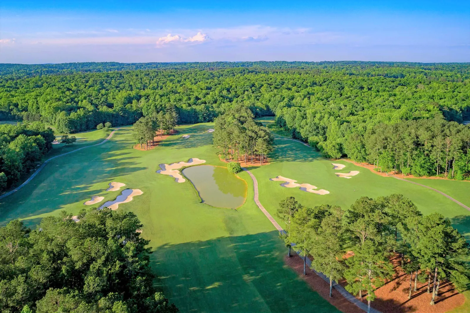 Aerial view of a golf course with fairways, sand bunkers, a water hazard, and surrounding forest under a clear sky.
