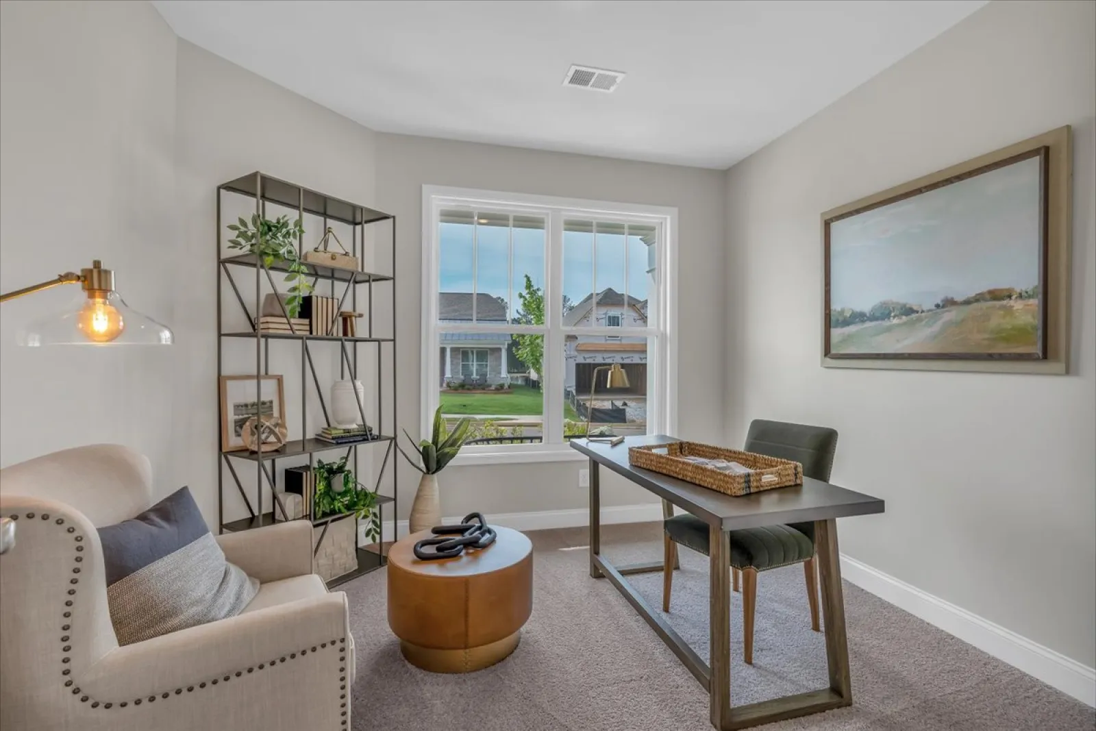 Cozy home office with beige armchair, modern desk, metal bookshelf, large window, and framed landscape painting.
