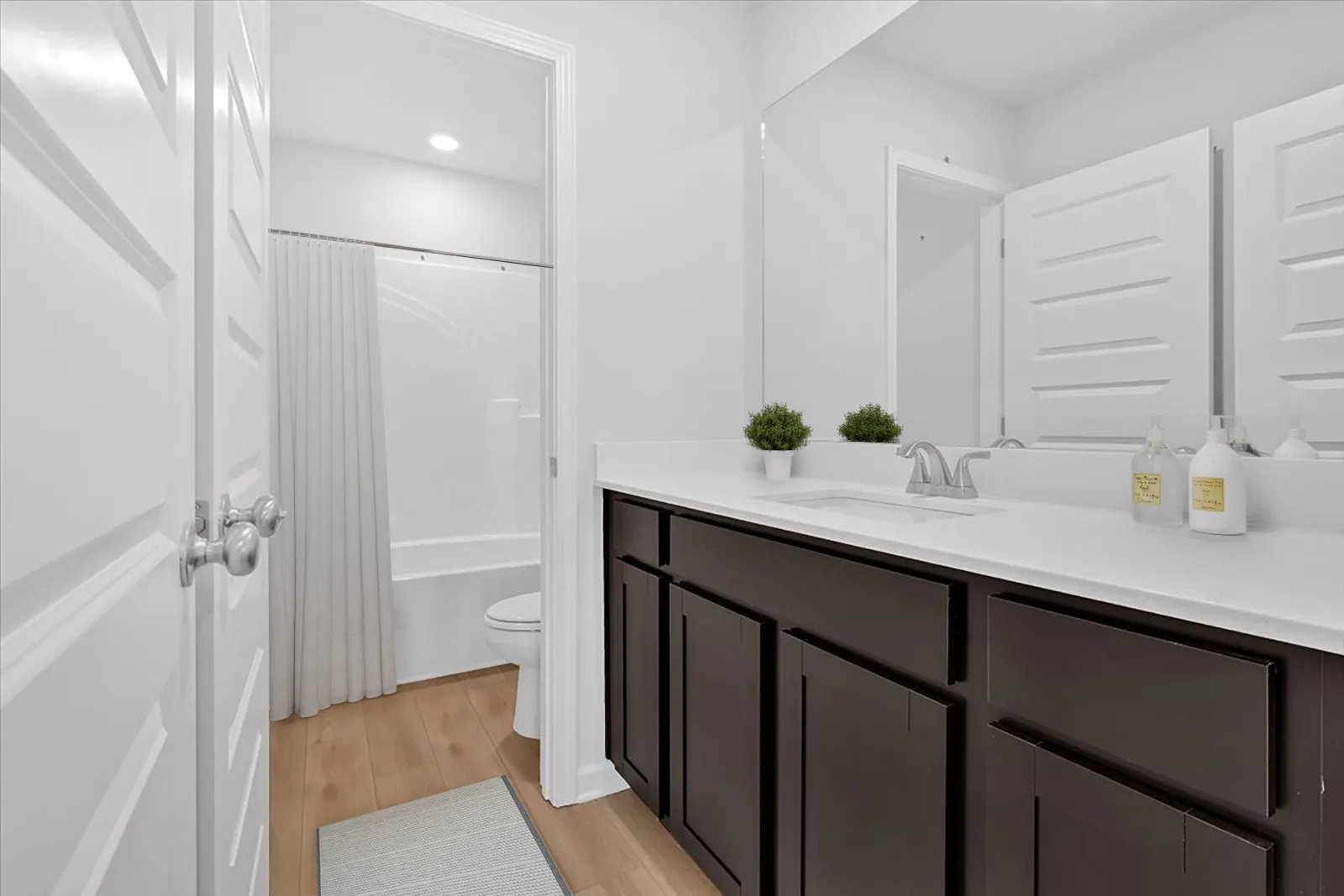 Modern bathroom with dark wood vanity, white countertop, large mirror, and white walls with hardwood floor.