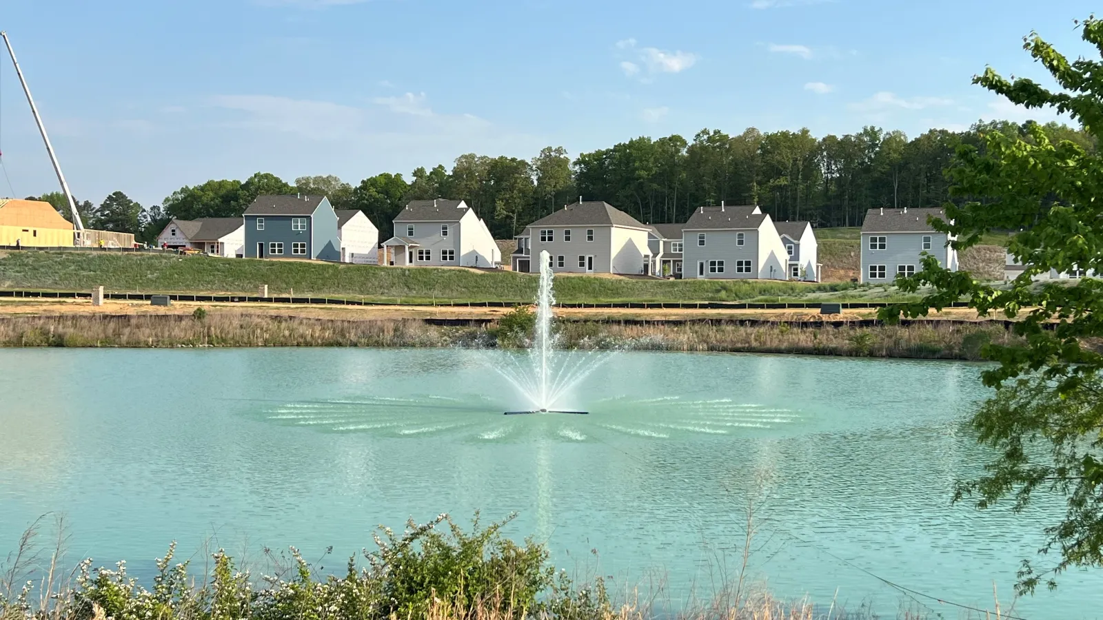 Residential houses behind a pond with a central water fountain under a clear blue sky.