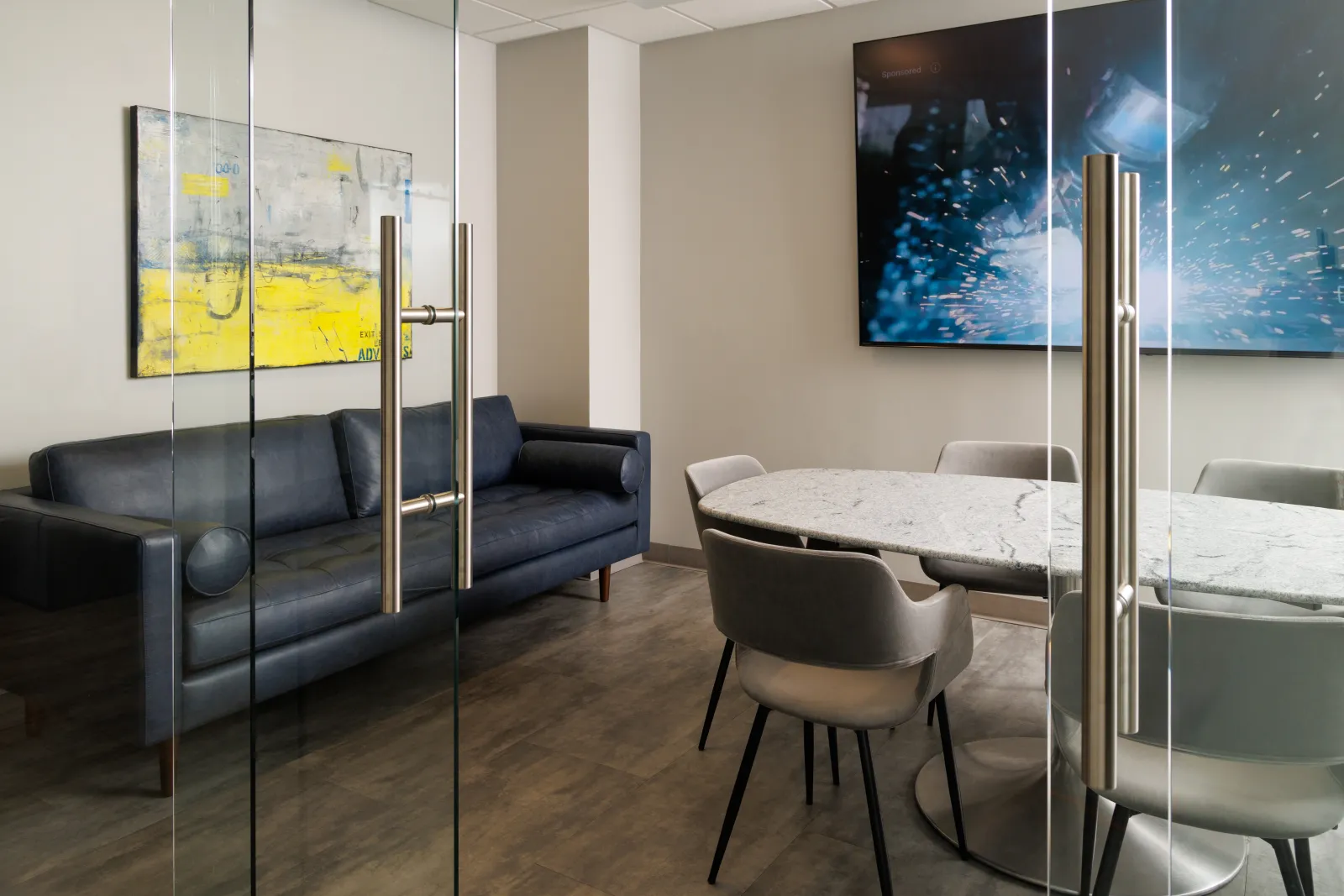Modern office meeting room with glass doors, marble table, grey chairs, black sofa, and abstract wall art.