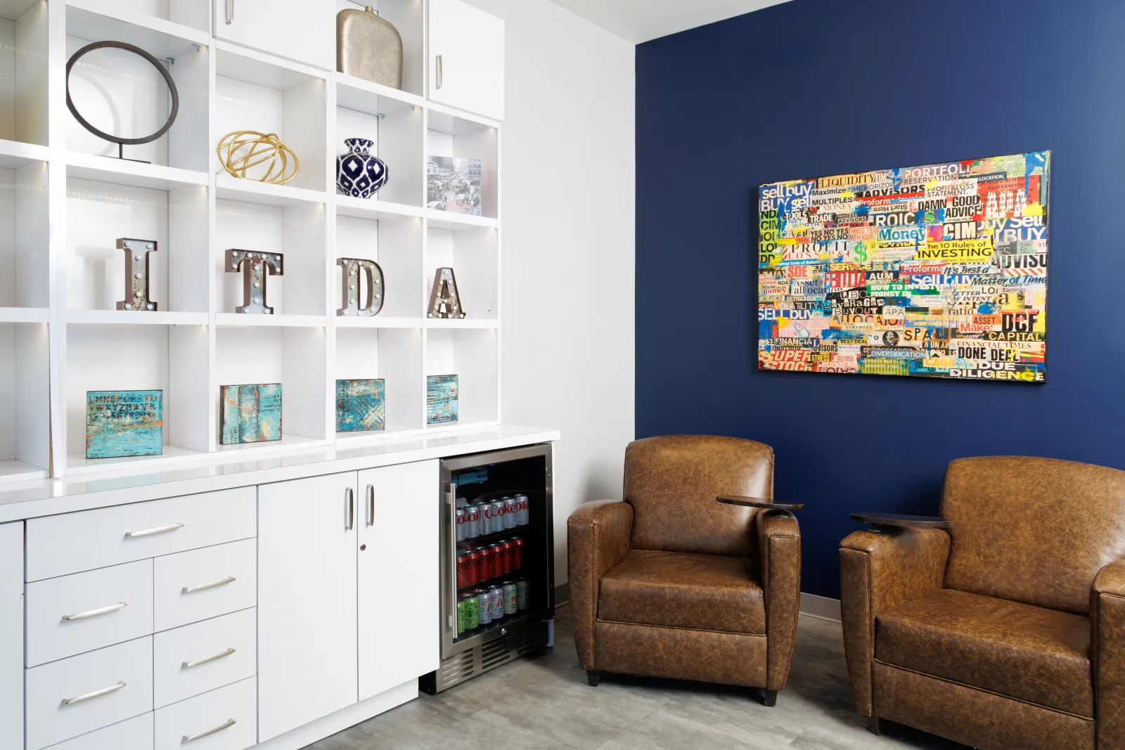 Modern office lounge with brown leather chairs, white shelving, mini fridge, and colorful wall art on blue wall.