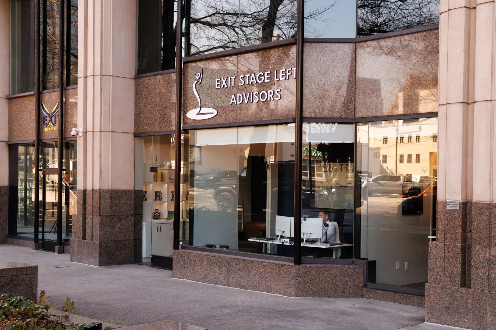 Office building exterior with large windows showing Exit Stage Left Advisors signage and a person working inside.