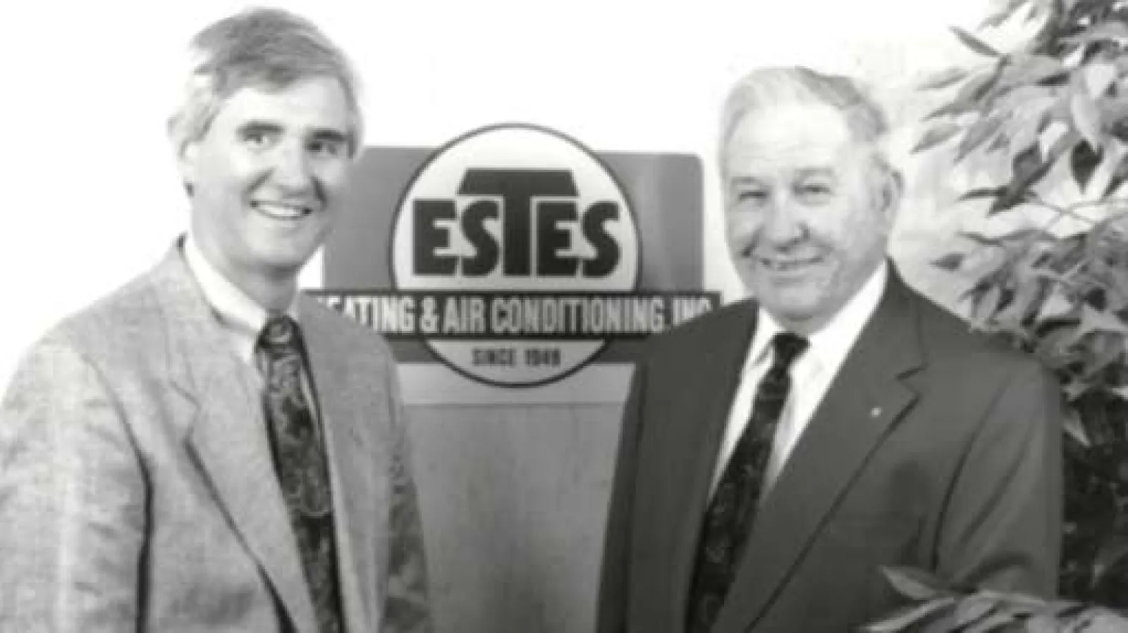 Mr Tommy and Nap Estes,  Founders of Estes Services smiling men in suits standing beside an Estes Heating & Air Conditioning sign in an office setting.