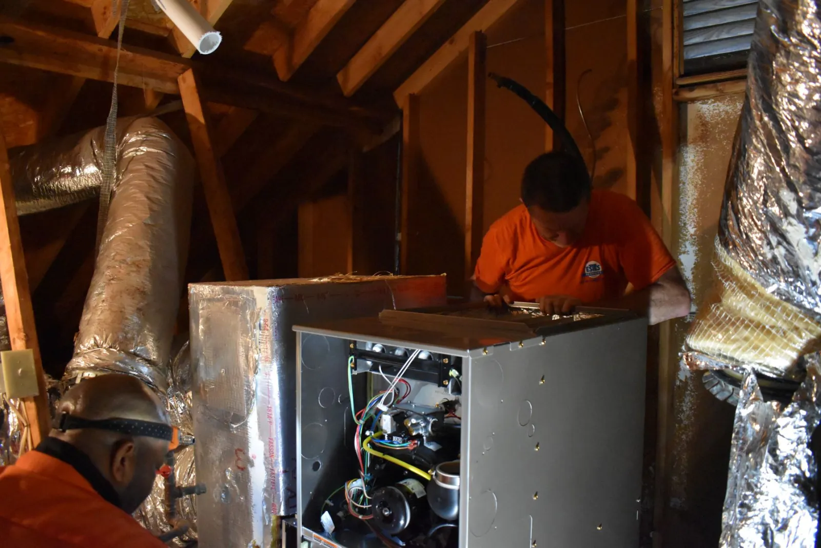 Two HVAC technicians working on installing or repairing a furnace in a dimly lit attic space