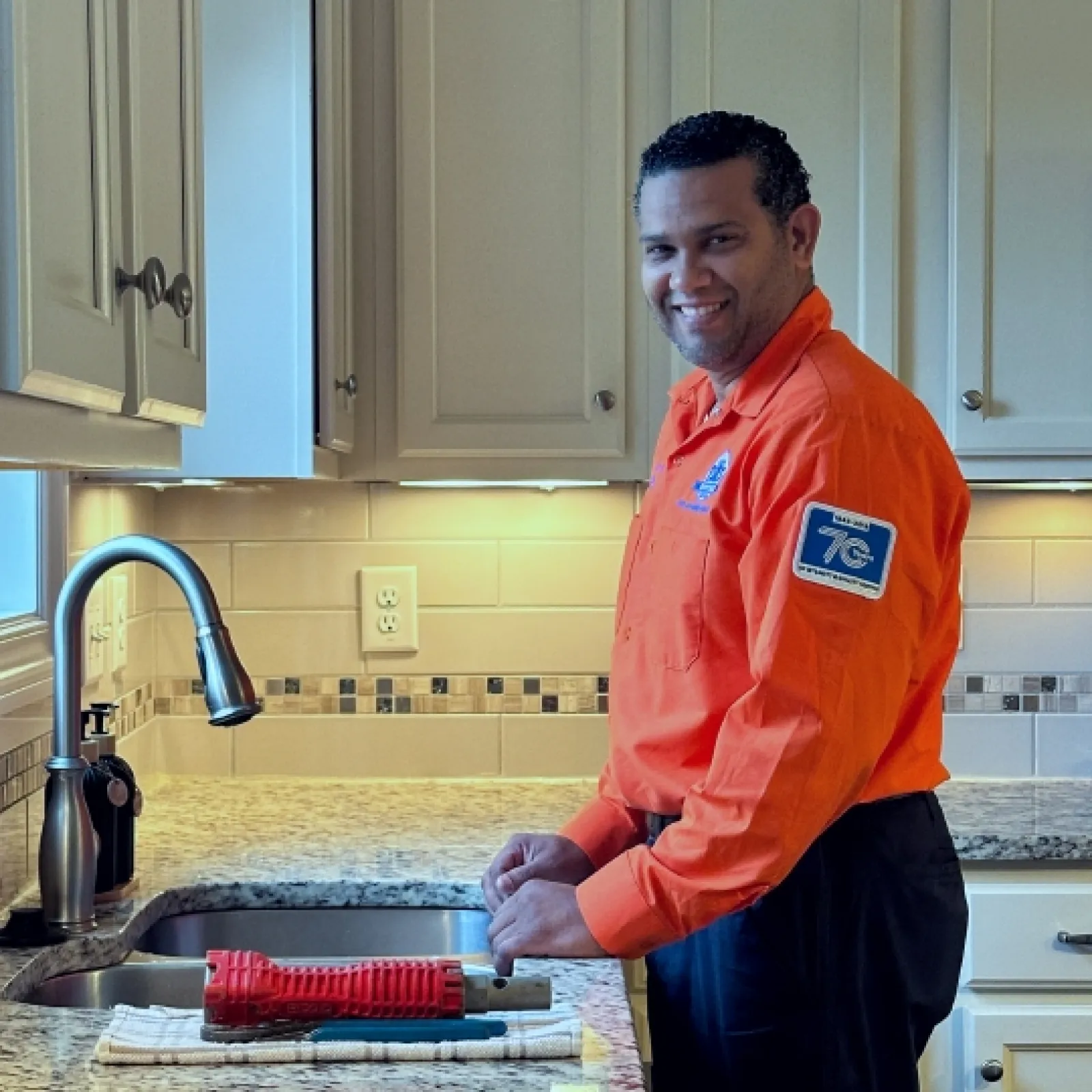 Smiling Estes plumber in orange uniform working at kitchen sink with tools on granite countertop under white cabinets.