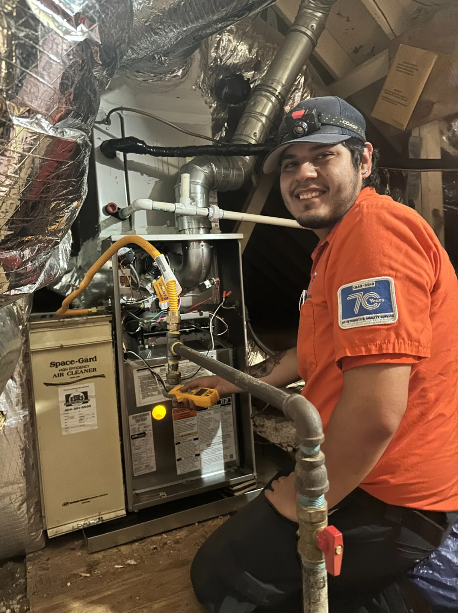 Estes Technician in orange uniform repairing a furnace in a basement with insulated pipes and tools around.
