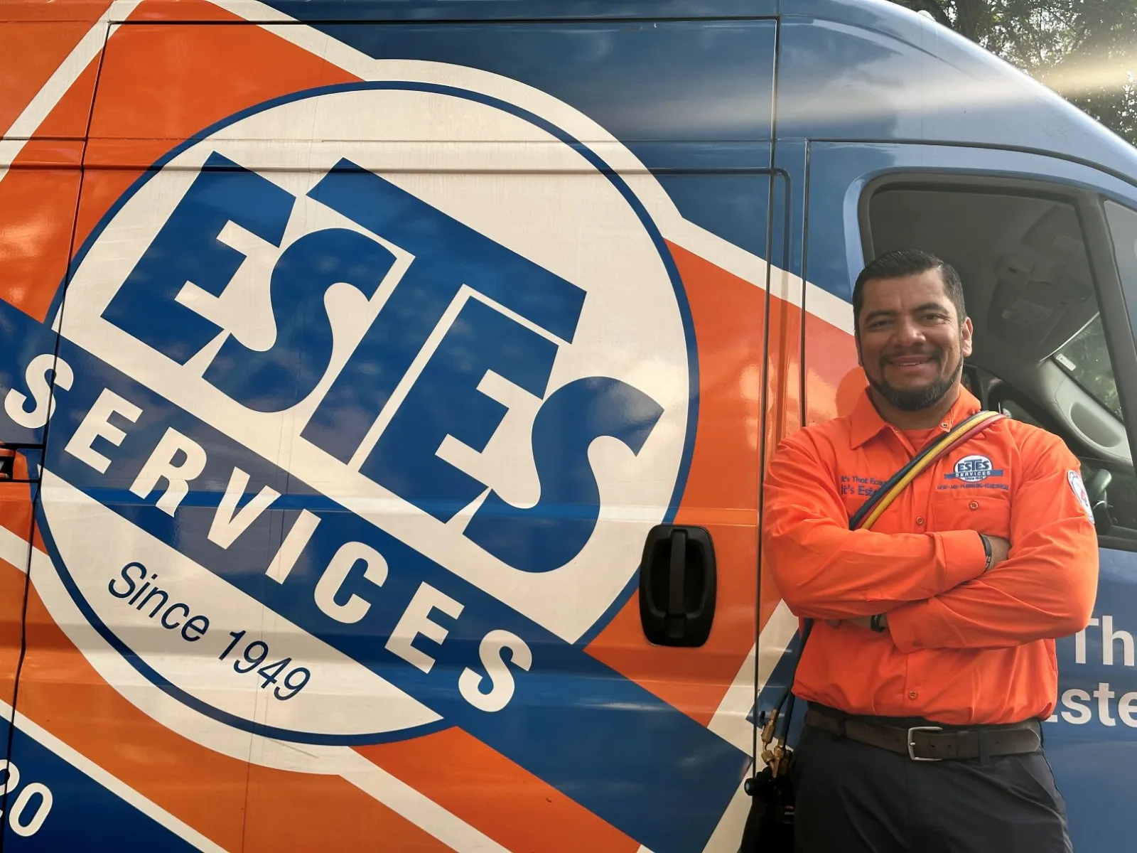 Smiling Estes Services HVAC Comfort Specialist  in orange shirt stands beside branded service Estes Services van 