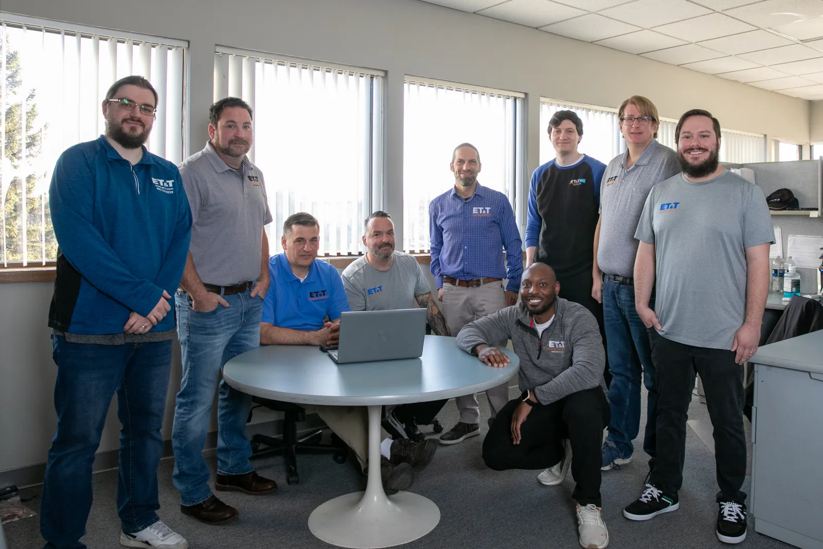 Group of nine diverse men posing around a table with a laptop in a bright office space with large windows.