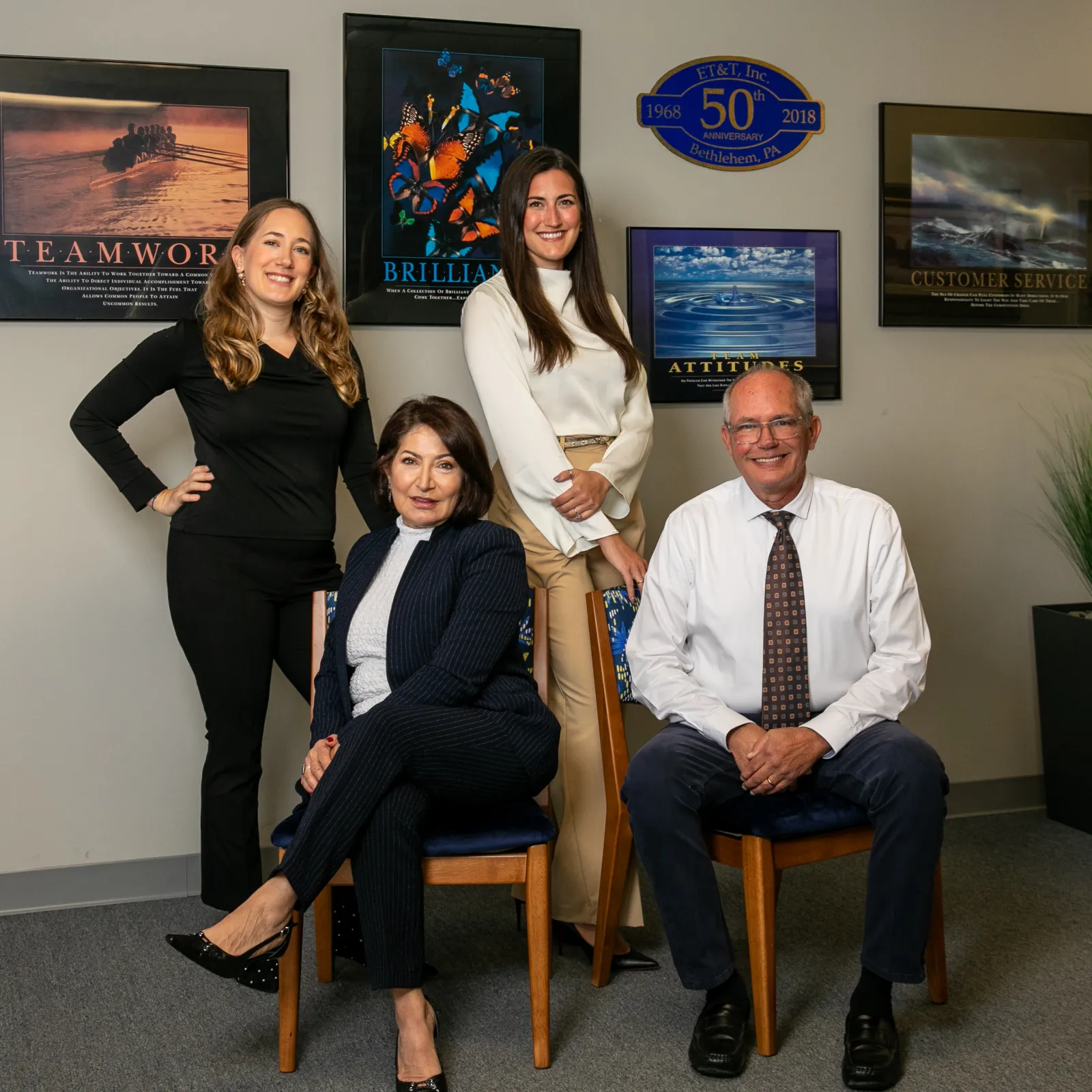 Four business professionals posing in office with motivational posters and anniversary plaque on the wall.