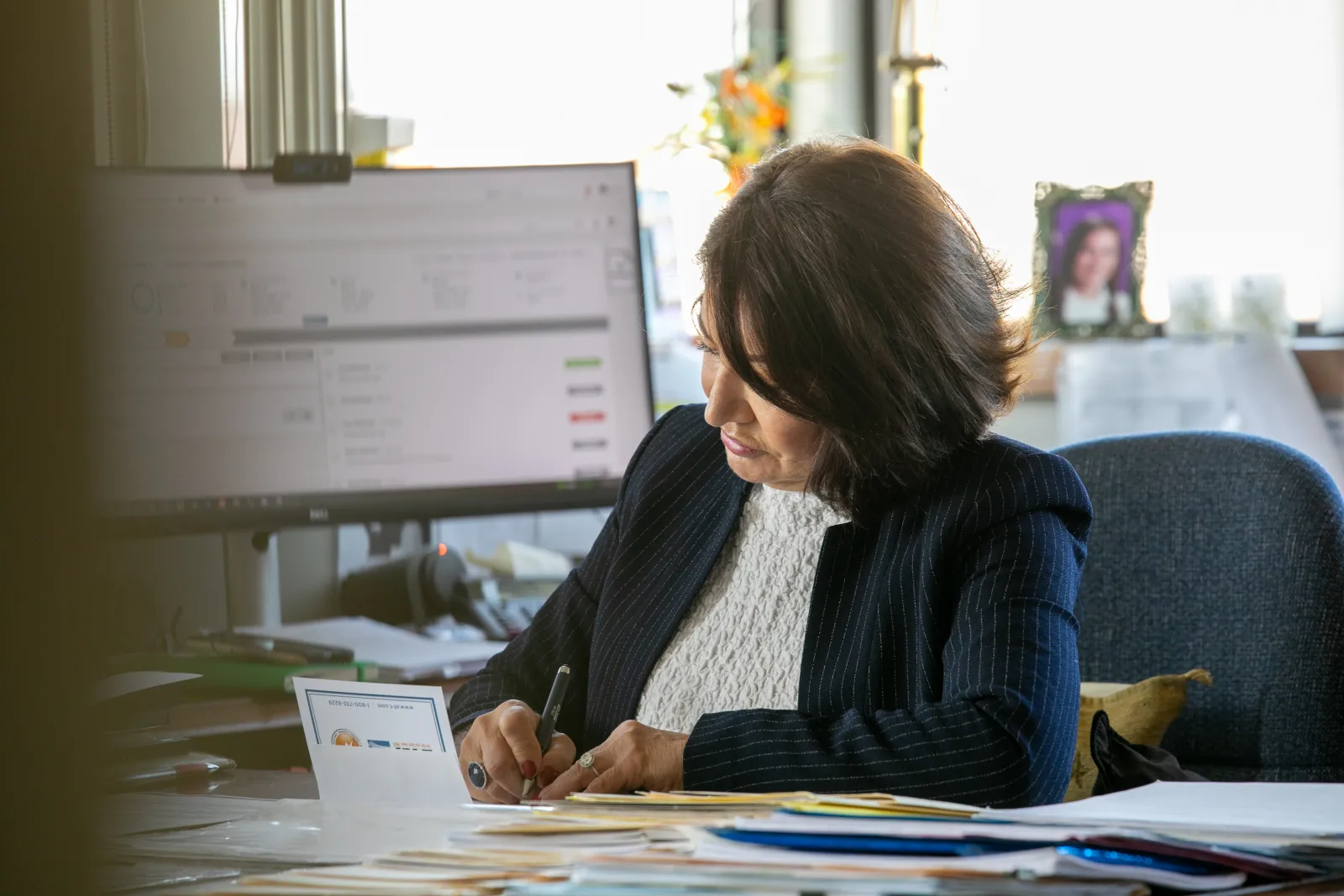 Professional woman in office writing notes with computer and paperwork visible in background.
