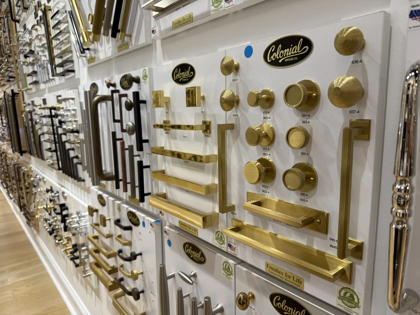 Wall display of various gold, black, and silver cabinet knobs and handles from Colonial Bronze Co.