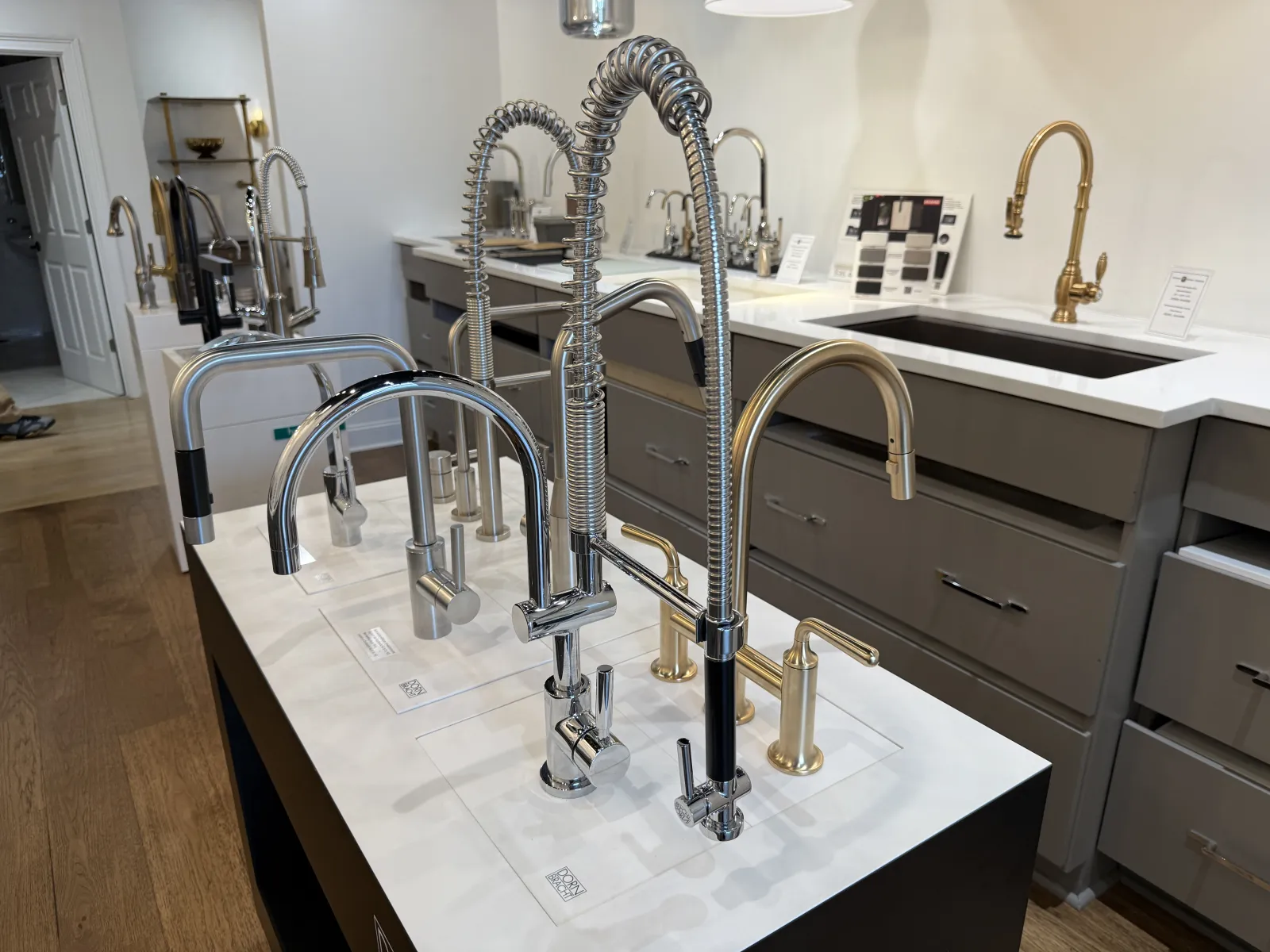 Modern kitchen faucets in various designs and finishes displayed on a white counter in a showroom.