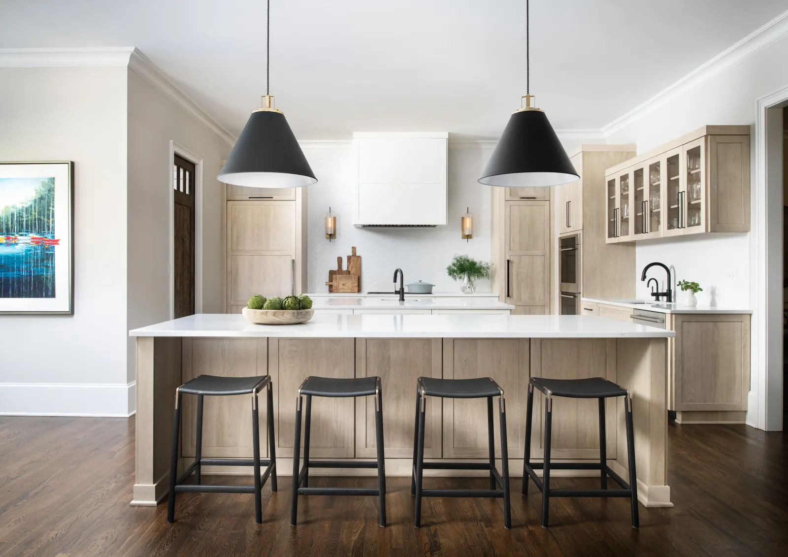 Modern kitchen with a large island, four black stools, pendant lights, and wood flooring under natural light.