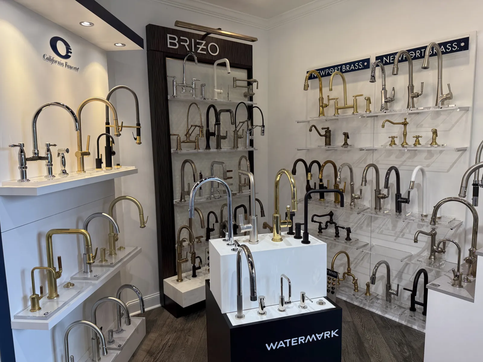 Display of various stylish kitchen and bathroom faucets from Brizo, Newport Brass, and Watermark brands in a showroom.