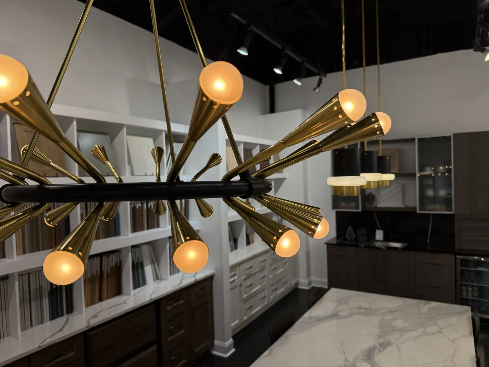 Modern kitchen with marble island, brass chandelier, dark cabinetry, and shelves filled with samples and books.