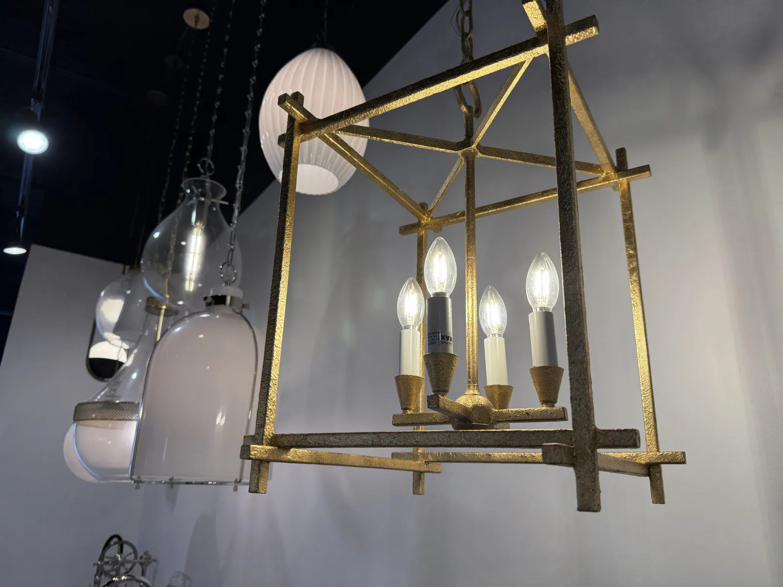 Gold metal chandelier with five candle-like bulbs hanging in modern showroom with other pendant lamps.