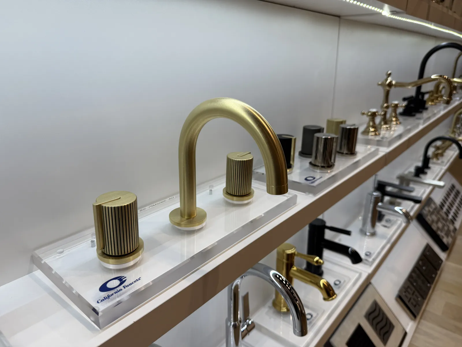 Display of various modern faucet designs including a prominent matte gold faucet with ribbed handles on a showroom shelf.