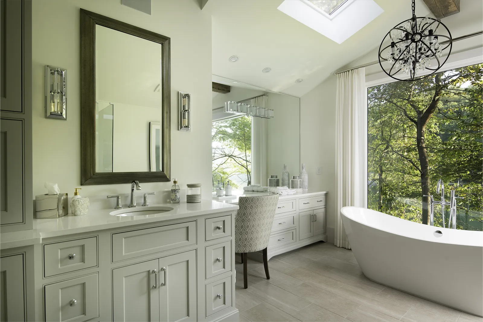 Bright modern bathroom with white vanity, freestanding tub, large mirror, and window view of trees.