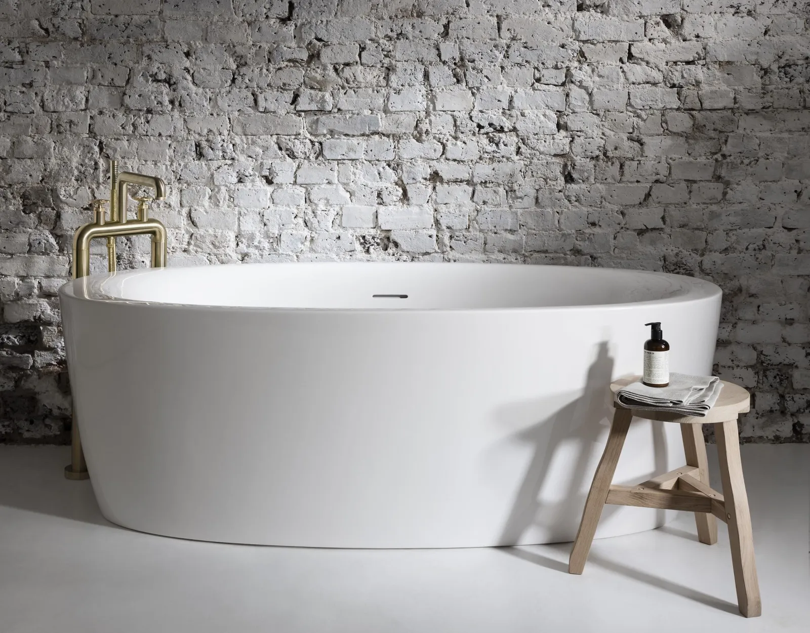 Modern freestanding bathtub beside a minimalist wooden stool against a rustic brick wall.